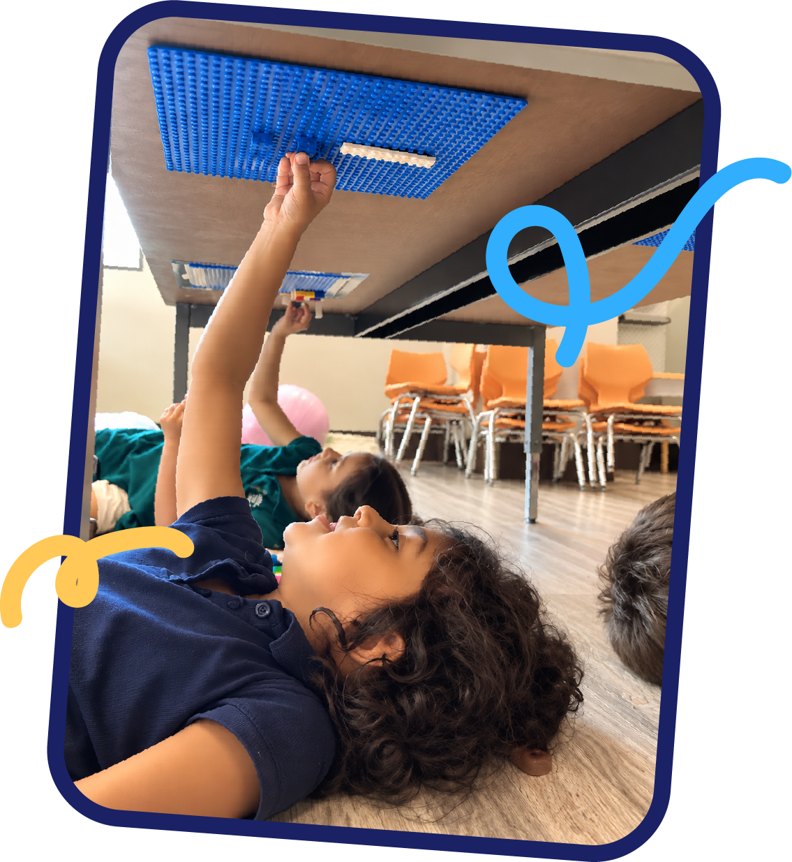 Children lying on the floor building with LEGO baseplates attached under a table, a creative STEM activity at STEM Bright Beginnings childcare and daycare in Leander, TX