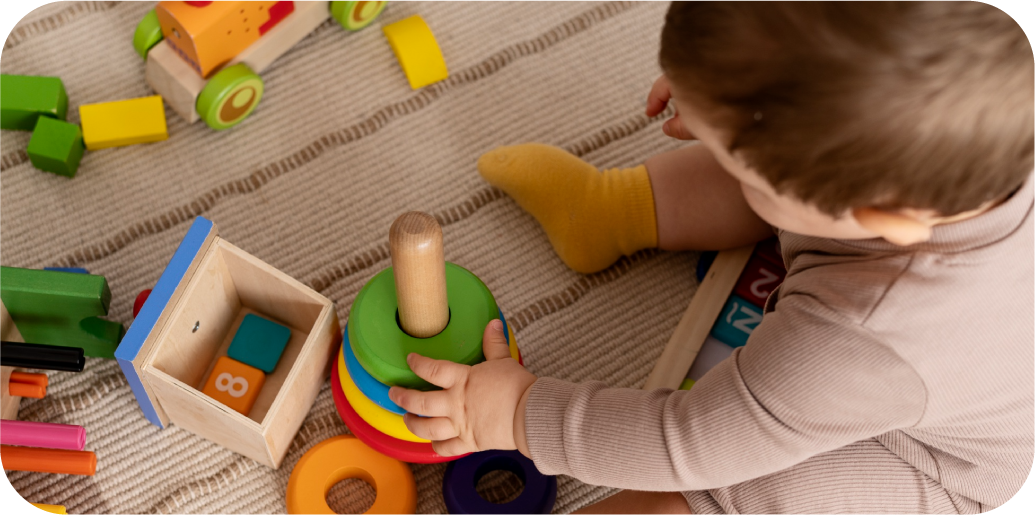 Infant playing with a wooden stacking ring toy surrounded by blocks and sensory toys in the Discoverers program at STEM Bright Beginnings in Leander, TX
